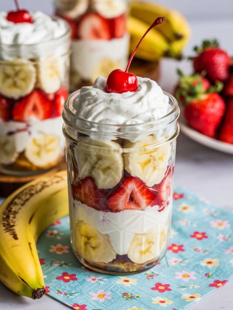 Mason jars filled with layers of banana, strawberry, whipped cream, and a cherry on top for an easy banana split dessert