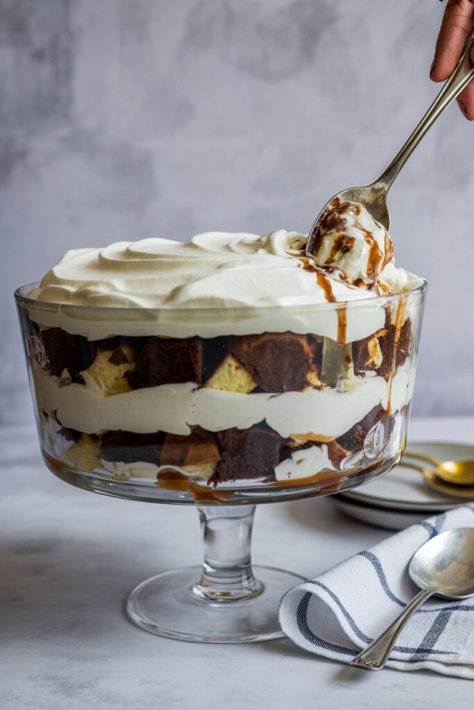 Layered chocolate trifle in a clear glass pedestal bowl, with whipped cream on top and a spoon lifting a scoop to show the layers of cake, cream, and chocolate.