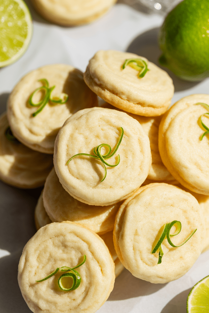 Real-life close-up image of soft lime sugar cookies, bright citrus desserts topped with lime zest and light glaze, arranged on a clean white surface in a modern kitchen with soft natural light.