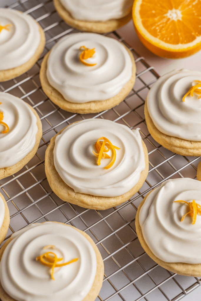 Real-life image of frosted orange cookies, soft citrus desserts topped with creamy orange icing and zest, arranged on a cooling rack in a bright white kitchen with soft natural window light and minimal props.