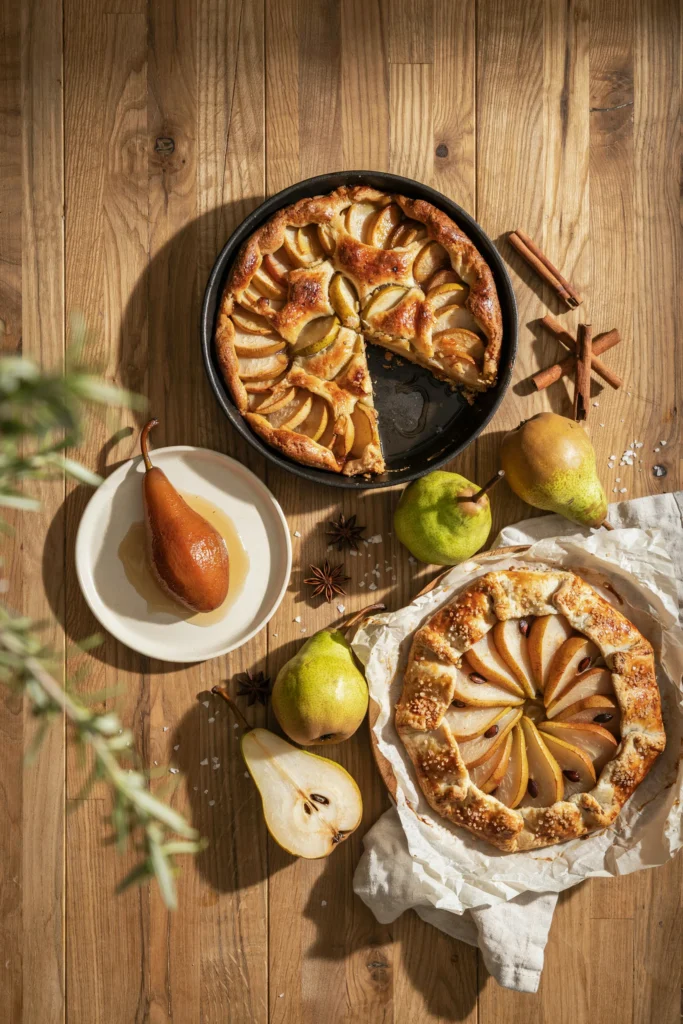 An overhead flat lay of three or four pear desserts — a golden cobbler, a glossy poached pear, and a rustic galette — surrounded by whole pears and a few cinnamon sticks, warm autumn light, no text overlay needed