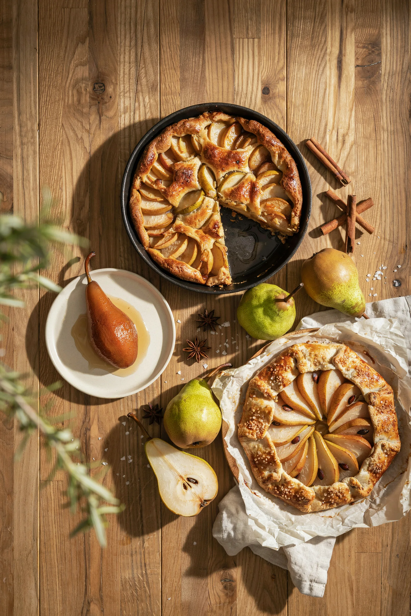 An overhead flat lay of three or four pear desserts — a golden cobbler, a glossy poached pear, and a rustic galette — surrounded by whole pears and a few cinnamon sticks, warm autumn light, no text overlay needed