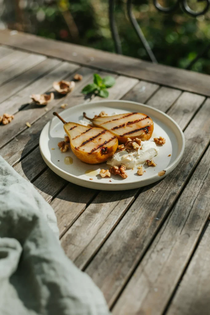 Halved pears with distinct char grill marks, served with a swirl of mascarpone and scattered toasted walnut pieces, on an outdoor table setting