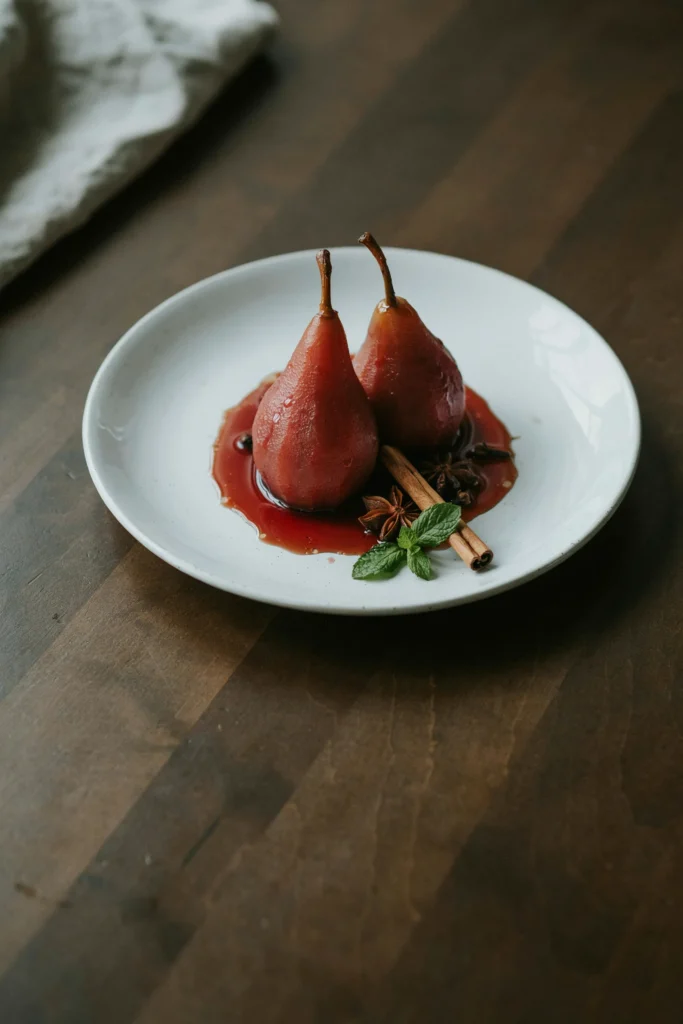 Whole pears poached to a deep ruby color in spiced wine, standing upright on a white plate with a pool of reduced wine syrup, garnished with a cinnamon stick