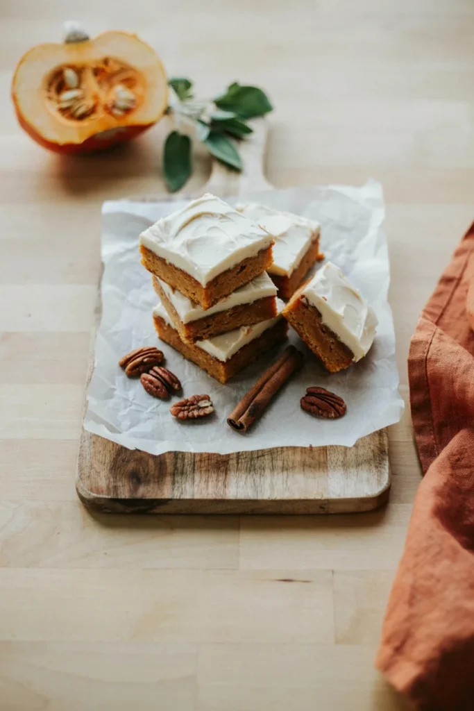 A stack of thick, soft pumpkin bars topped with swirled cream cheese frosting, cut into neat squares on parchment paper.