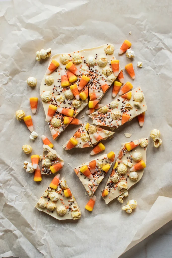 A broken slab of white chocolate popcorn bark studded with candy corn and orange and brown sprinkles, pieces scattered on parchment.