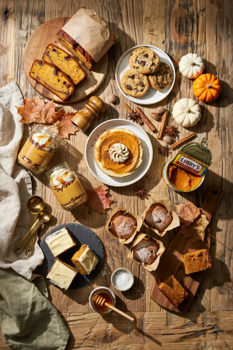 Overhead shot of a rustic wood table with a sliced pumpkin loaf, a stack of pumpkin cookies, and a jar of pumpkin dip, scattered cinnamon sticks and a few mini pumpkins around the edges, warm golden light, cozy fall mood