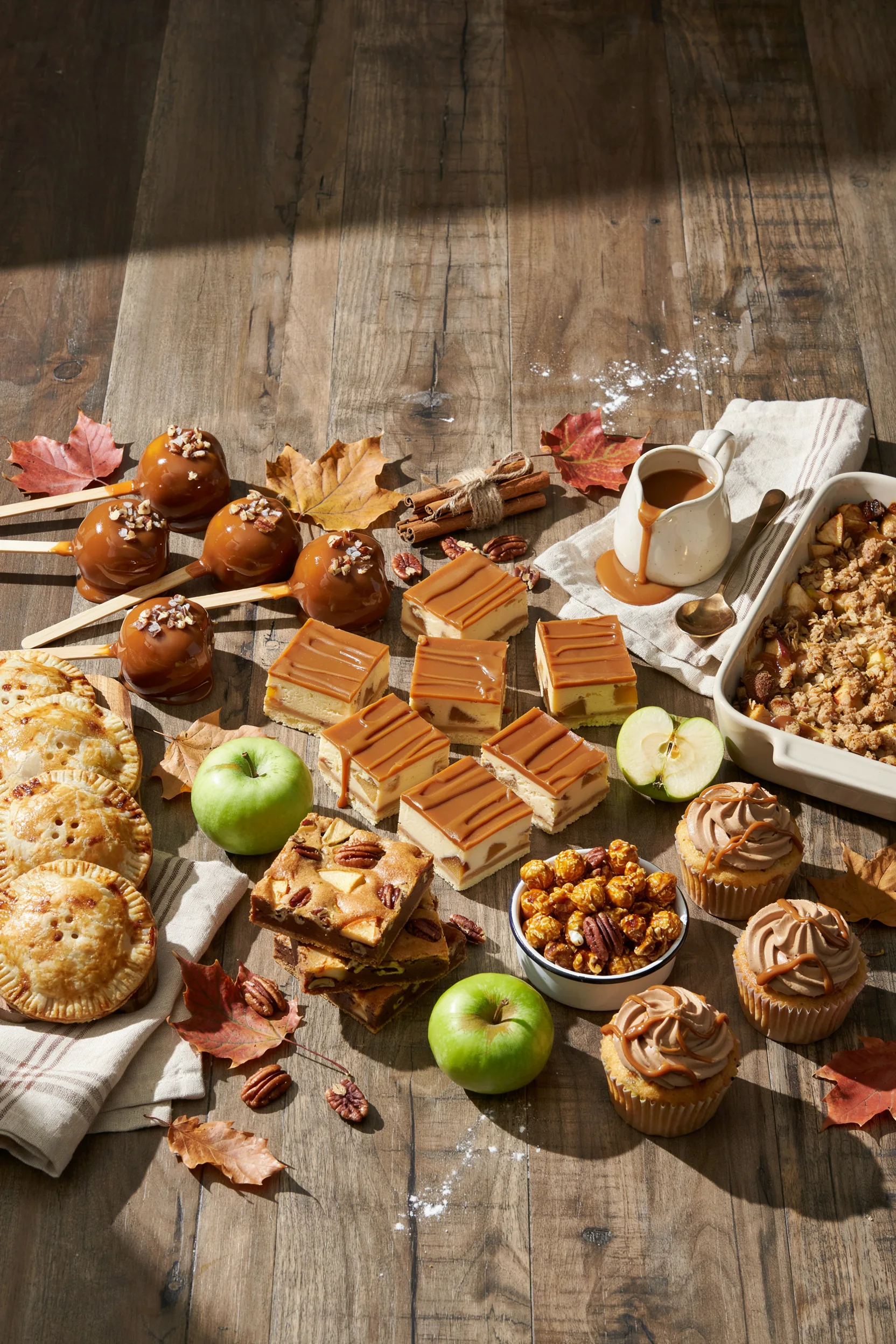 : An overhead flat-lay of several caramel apple desserts arranged together — dipped caramel apples, a slice of caramel apple cheesecake bar, and a bowl of caramel apple crisp — on a rustic wood table with scattered cinnamon sticks and a small pitcher of caramel sauce. Warm, golden lighting, cozy fall mood