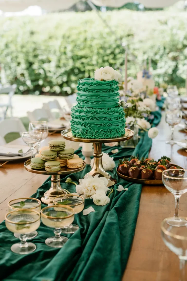 A styled dessert table shot from a slight overhead angle, deep emerald green desserts and linens layered with gold cake stands and white florals, soft natural light