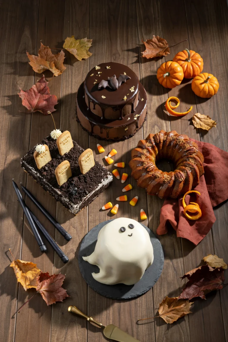 An overhead flat-lay of three or four finished Halloween cakes side by side — a black drip cake, an orange pumpkin bundt, and a graveyard dirt cake — on a dark wood table with scattered candy corn and mini pumpkins. Warm, moody lighting
