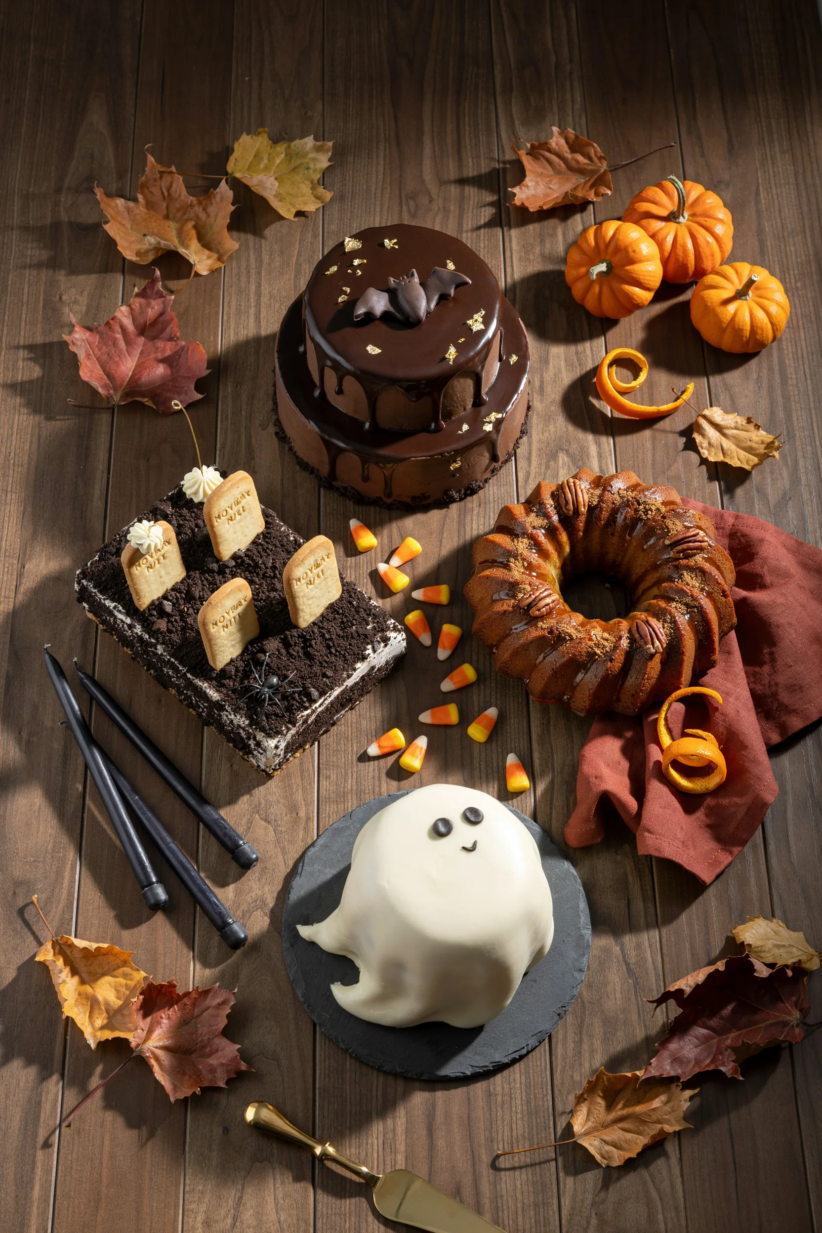 An overhead flat-lay of three or four finished Halloween cakes side by side — a black drip cake, an orange pumpkin bundt, and a graveyard dirt cake — on a dark wood table with scattered candy corn and mini pumpkins. Warm, moody lighting
