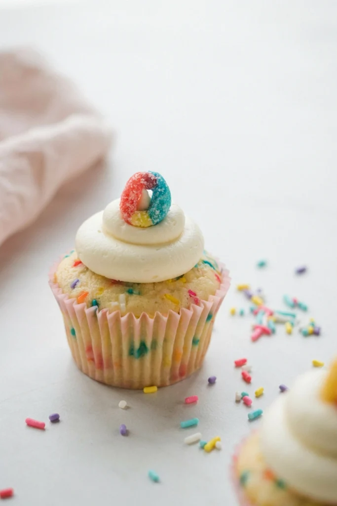 Funfetti cupcake with rainbow sprinkles topped with a white frosting swirl and a colorful alphabet cereal letter