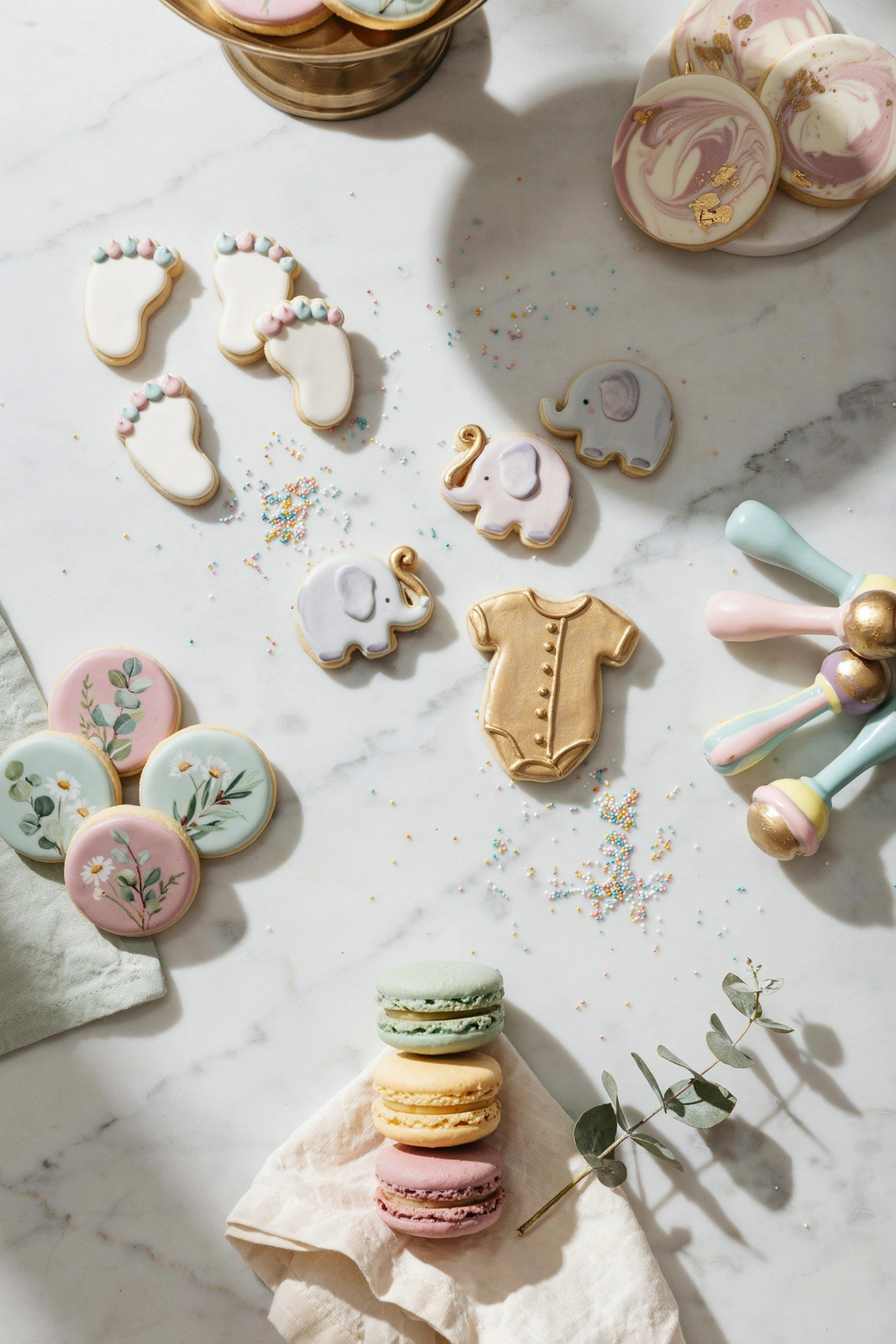 Overhead flat lay of an assortment of decorated baby shower cookies — pastel buttercream, gold-dusted onesies, watercolor animals — arranged on a marble surface with a few loose sprinkles scattered around.