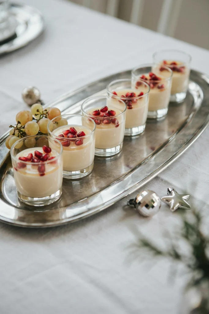 Small glass cups of pale gold panna cotta topped with a thin gelée layer and a few pomegranate arils, arranged on a silver tray.