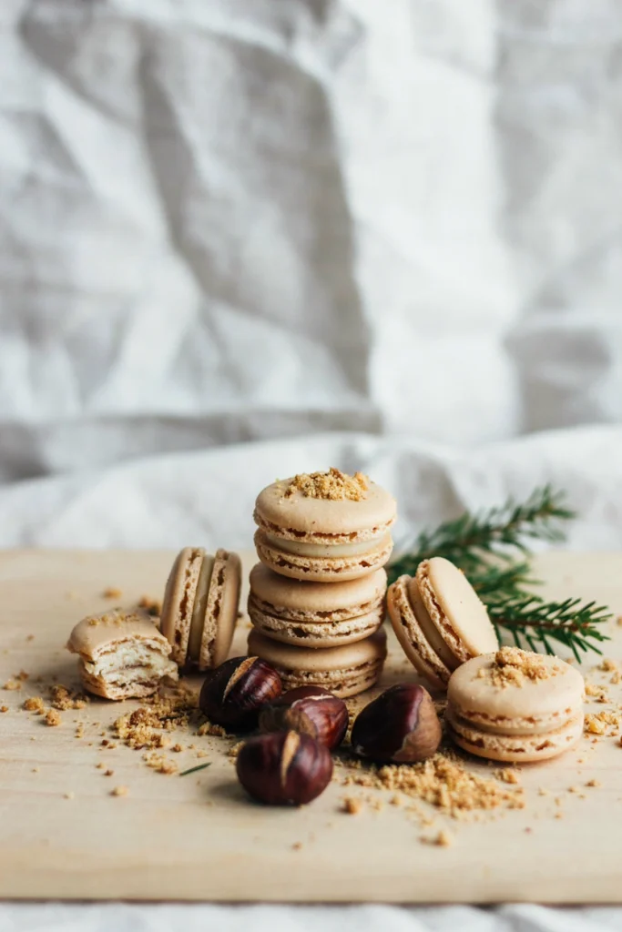 Pale beige macaron shells with a crushed praline crumble pressed into the top edge, filled with chestnut cream and stacked on a wooden board.